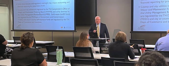 A presenter speaks to attendees in a classroom while slides about utility accounting and financial reporting are displayed on two screens.