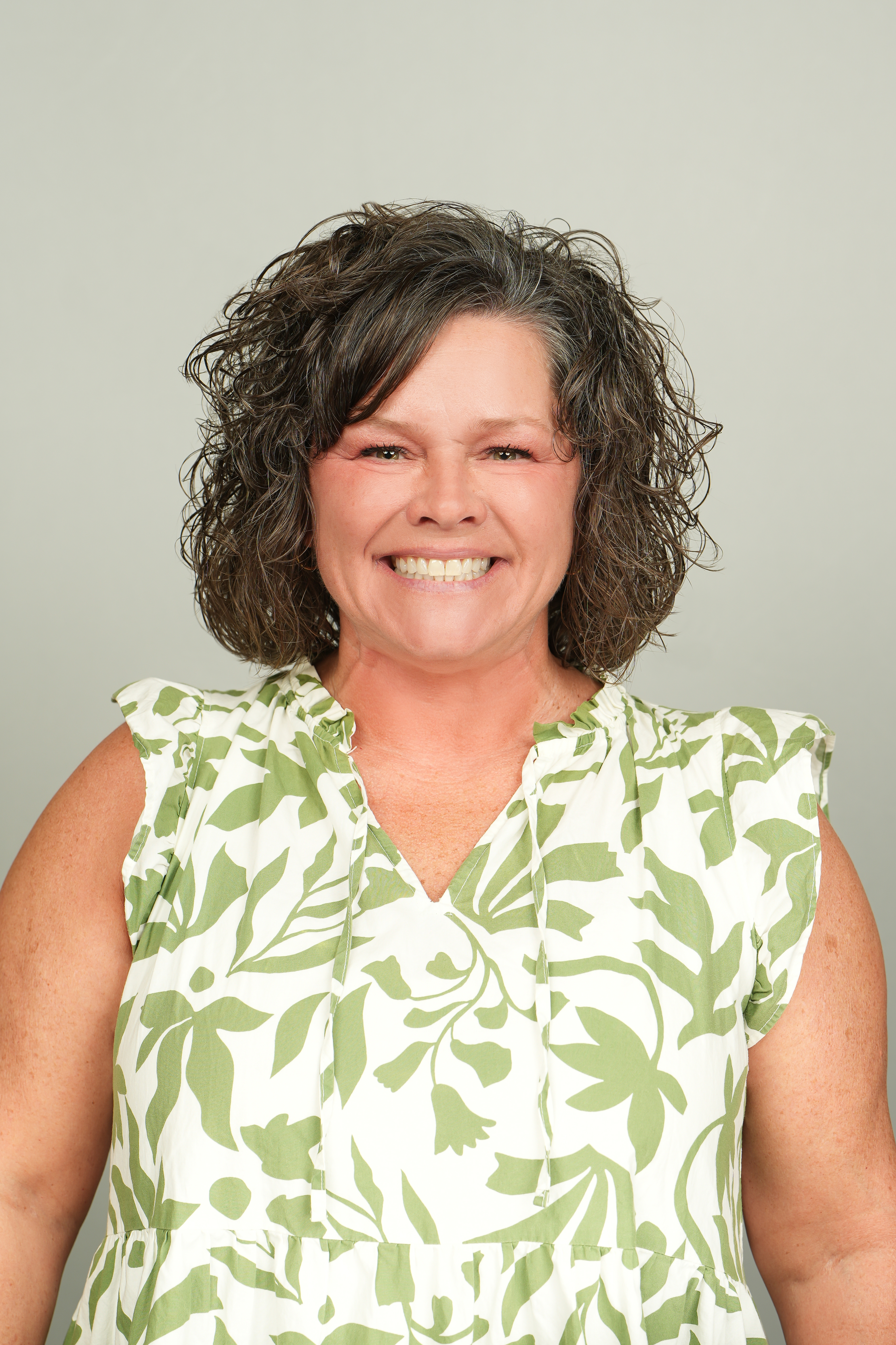 Professional headshot of a smiling person with short, curly brown hair, wearing a green-and-white patterned top.