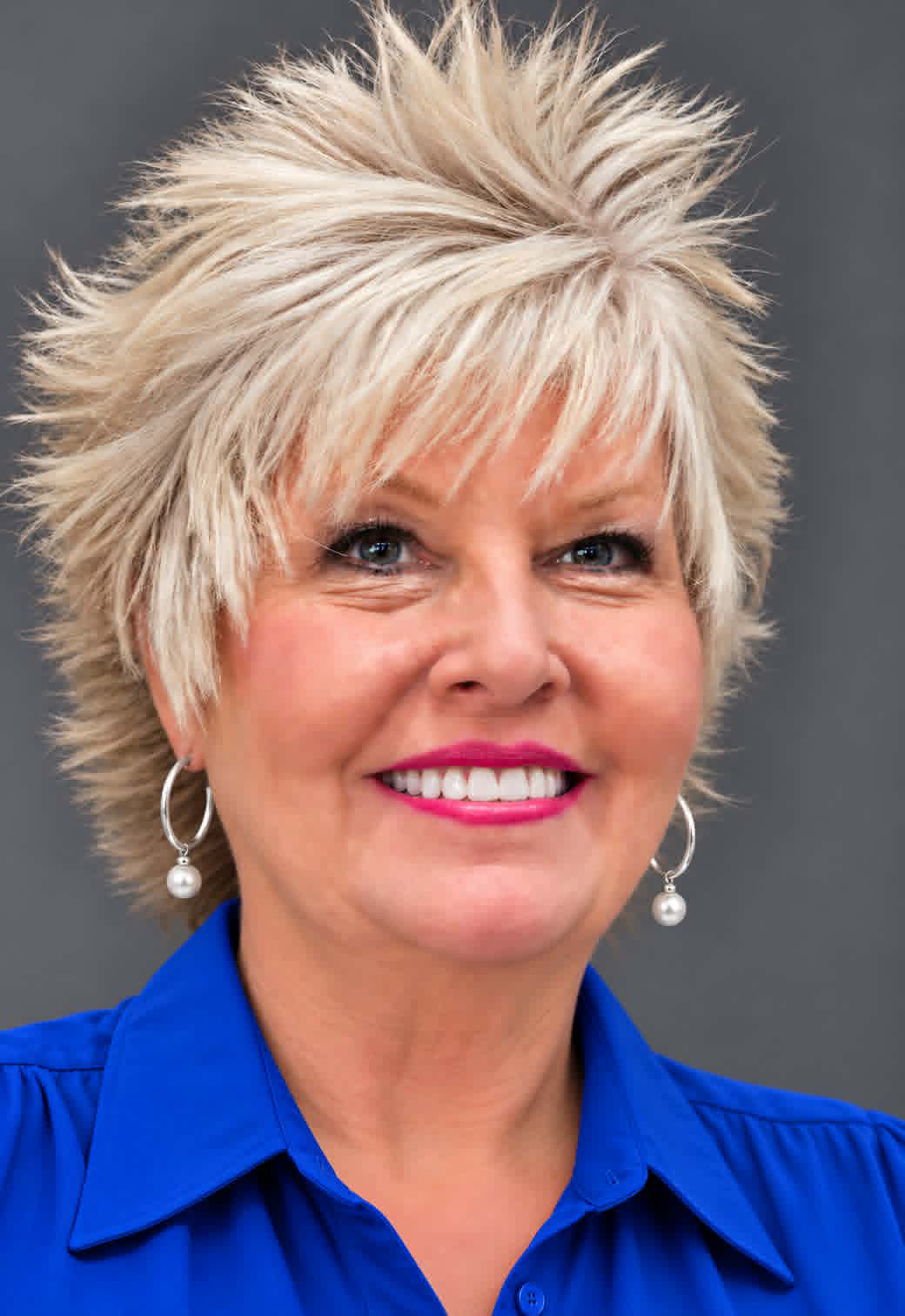 Woman with short, spiky blonde hair wearing a bright blue collared shirt and silver hoop earrings against a gray background.
