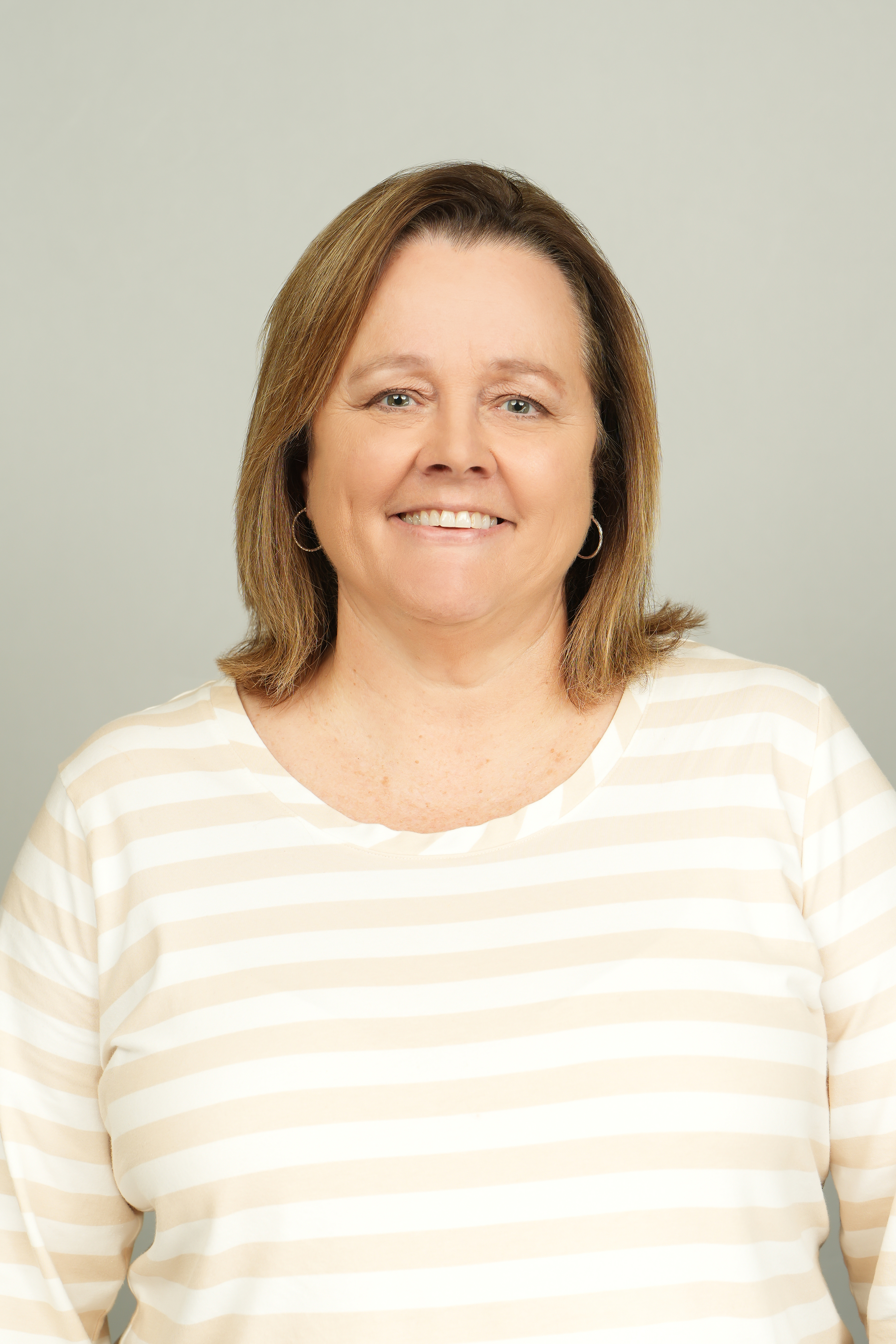 Professional headshot of a smiling person with shoulder-length brown hair, wearing a cream-and-white striped top.