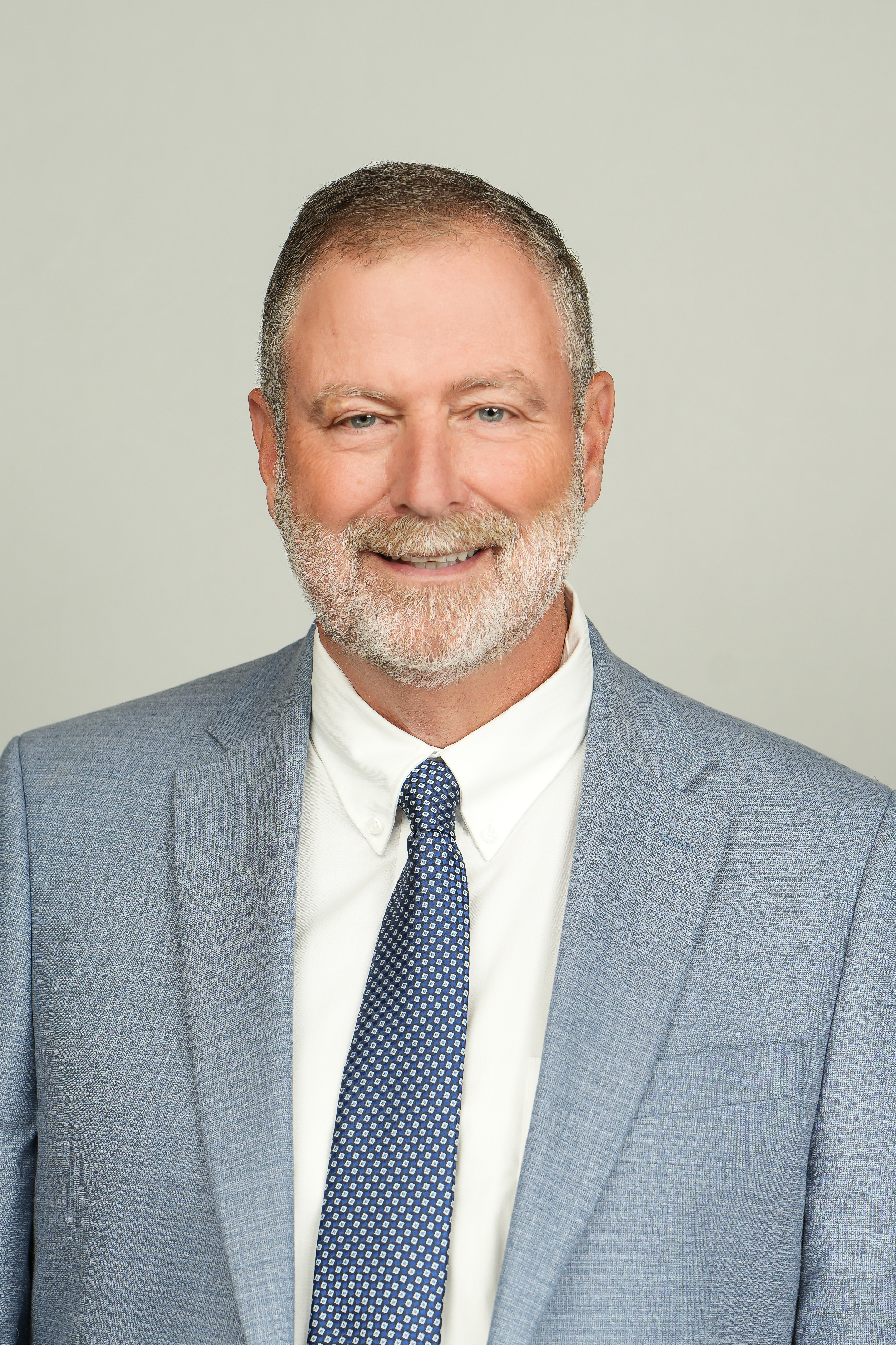 Professional headshot of a smiling person with short gray hair and a white beard, wearing a blue suit and patterned tie.