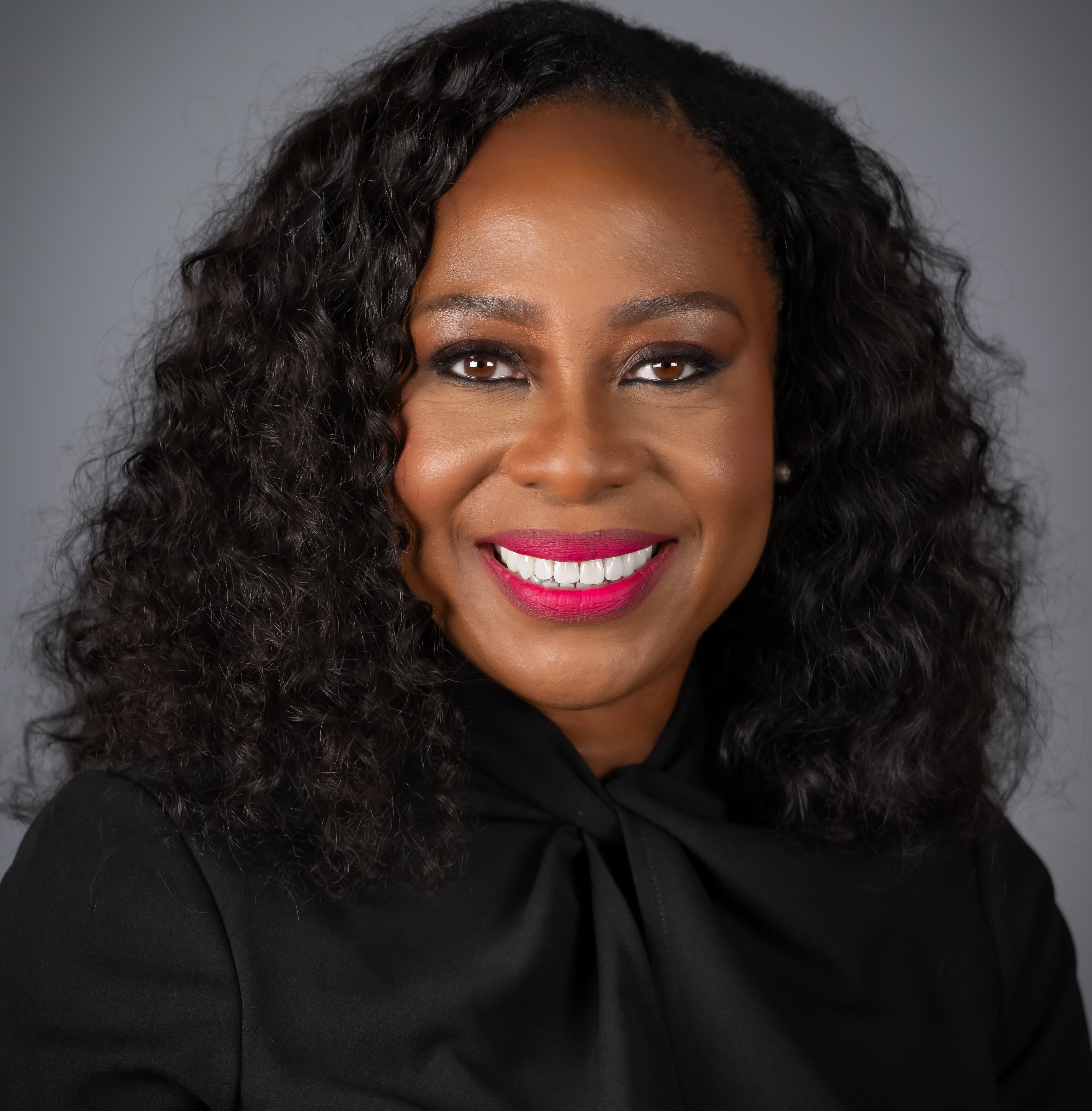 A woman with long curly dark hair smiles at the camera, wearing a black top against a gray background.