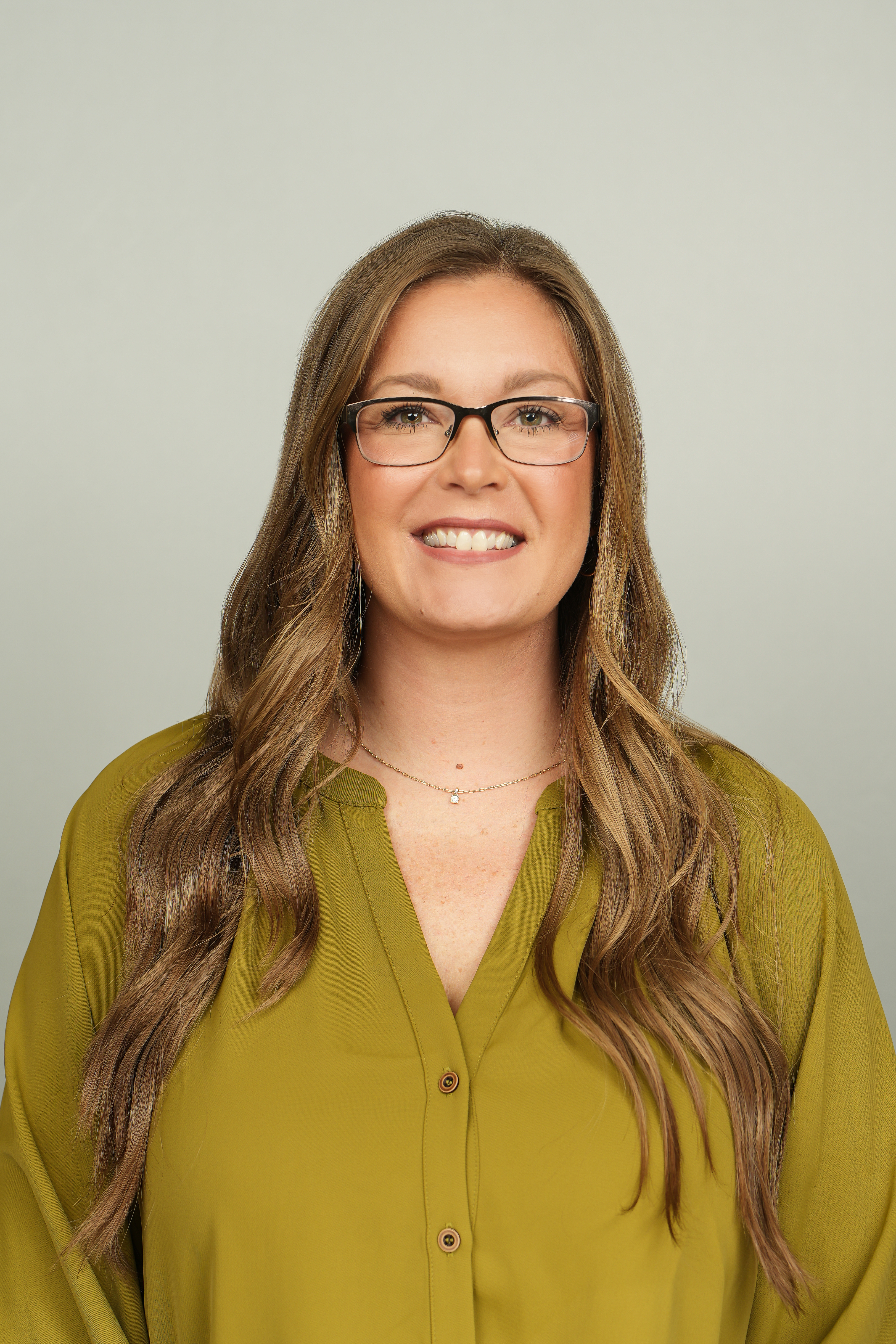Smiling woman with long brown hair and glasses wearing an olive green blouse against a light gray background.