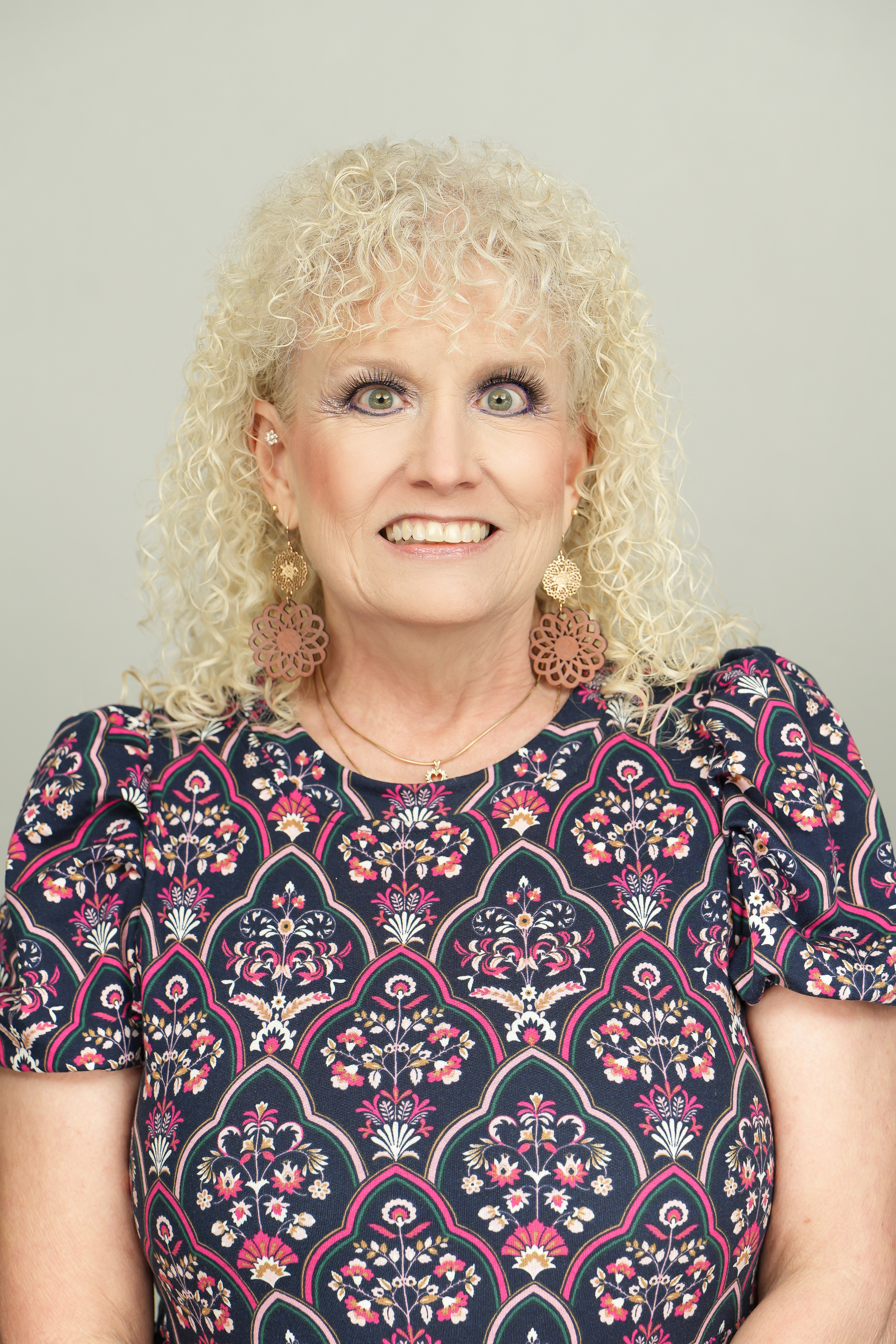 Professional headshot of a smiling person with curly blond hair, wearing a dark floral-patterned top and large earrings.