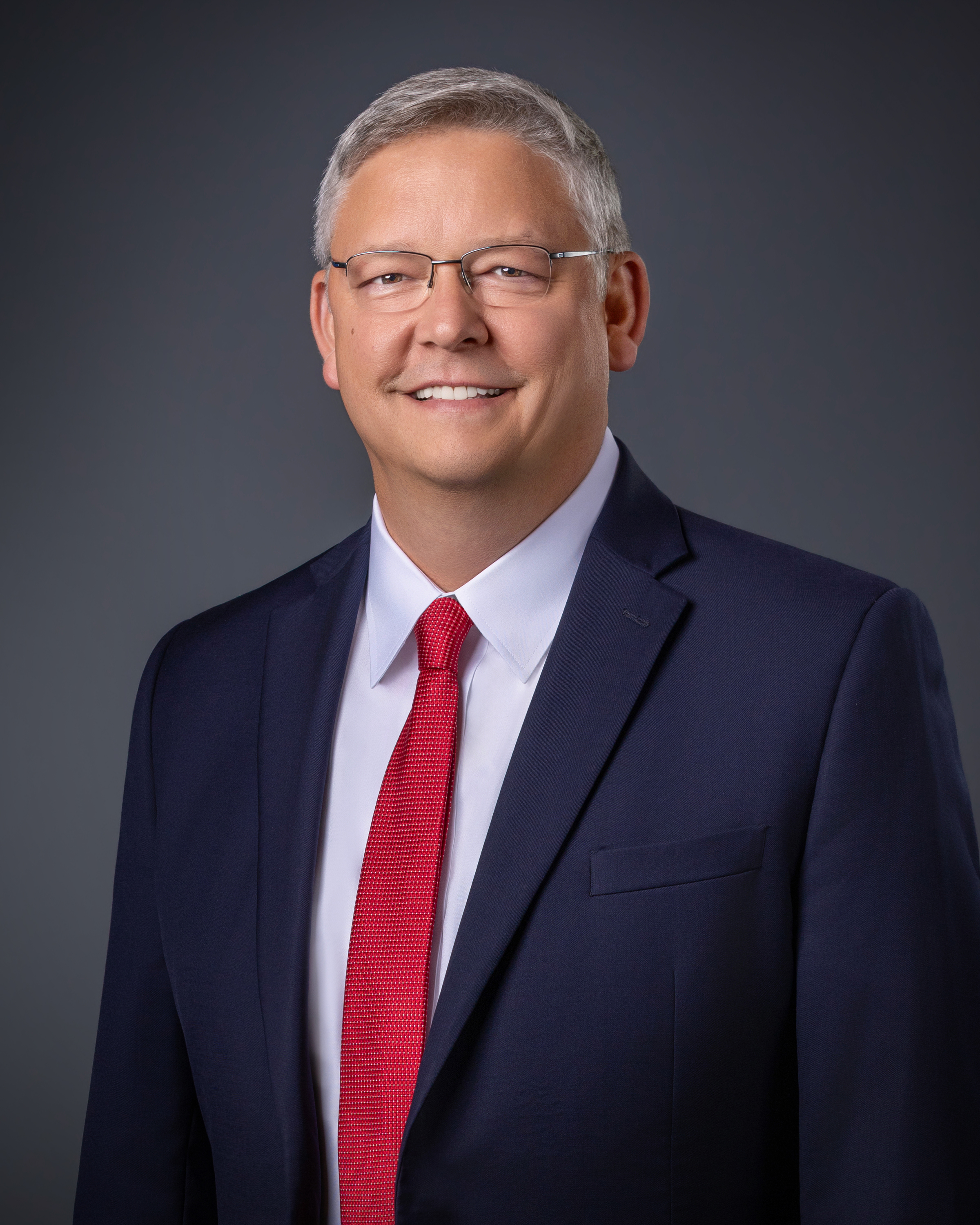 Professional headshot of Tipton County Executive Billy Daugherty wearing glasses, a navy suit, and a red tie.