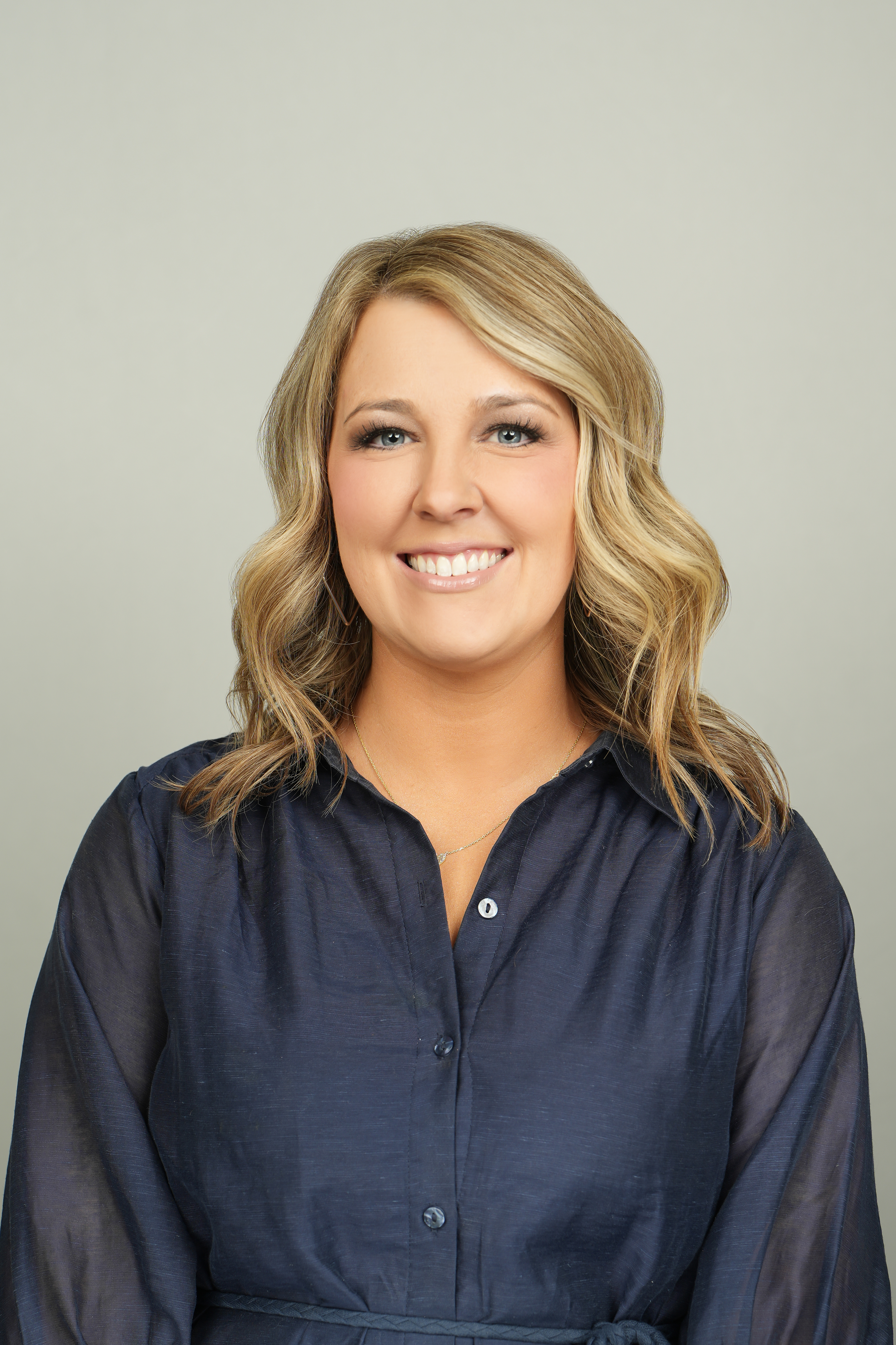 Professional headshot of a smiling person with shoulder-length blond hair, wearing a navy blouse.