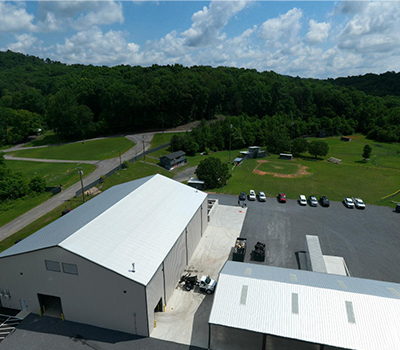 Aerial view of a white industrial facility with a parking lot, open grassy areas, and surrounding forest under a partly cloudy sky.